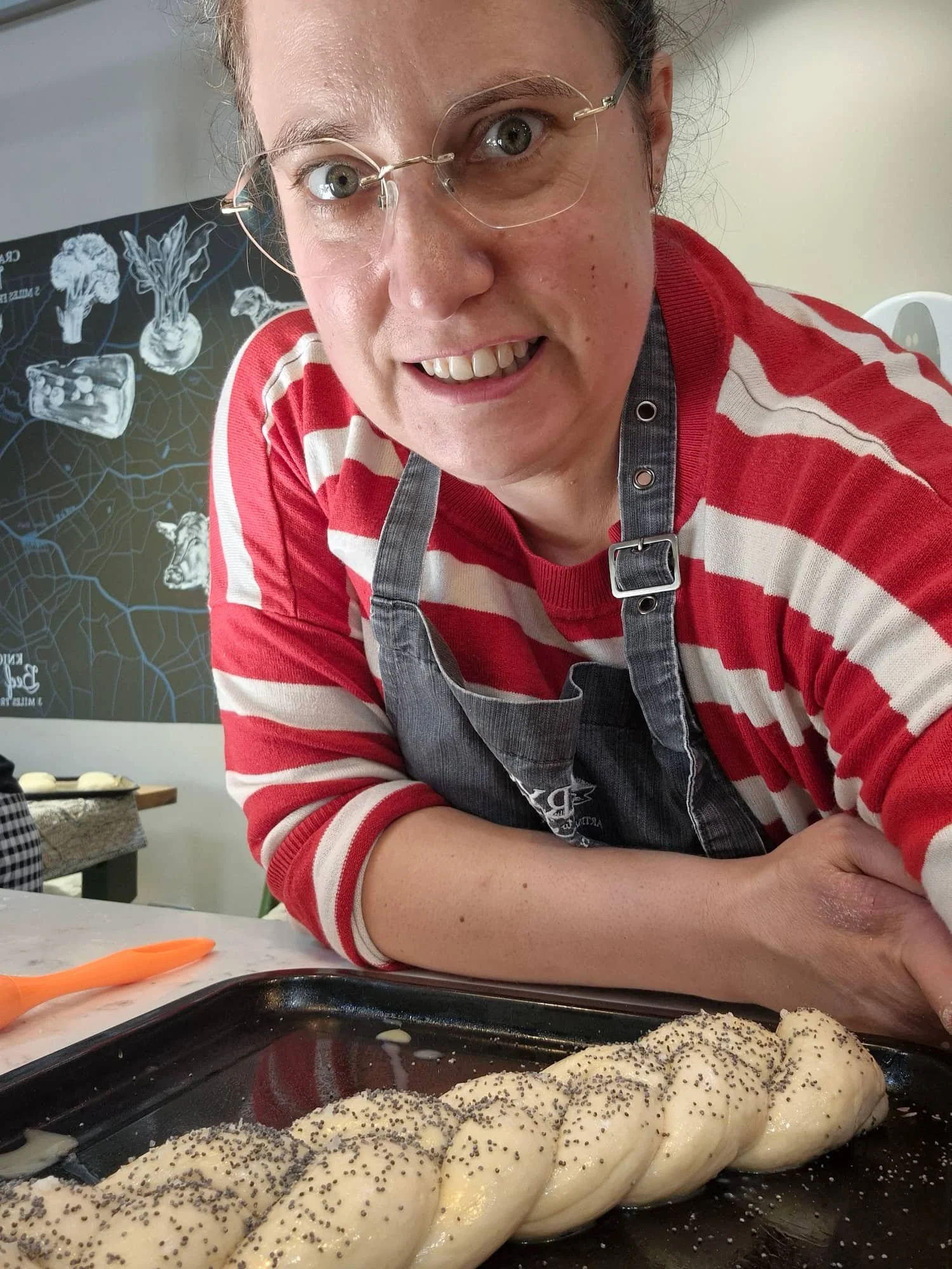 Woman in a striped jumper and grey apron smiles over a raw, poppy-seed-topped plaited bread loaf on a baking tray at a bakery in Eccleshall.