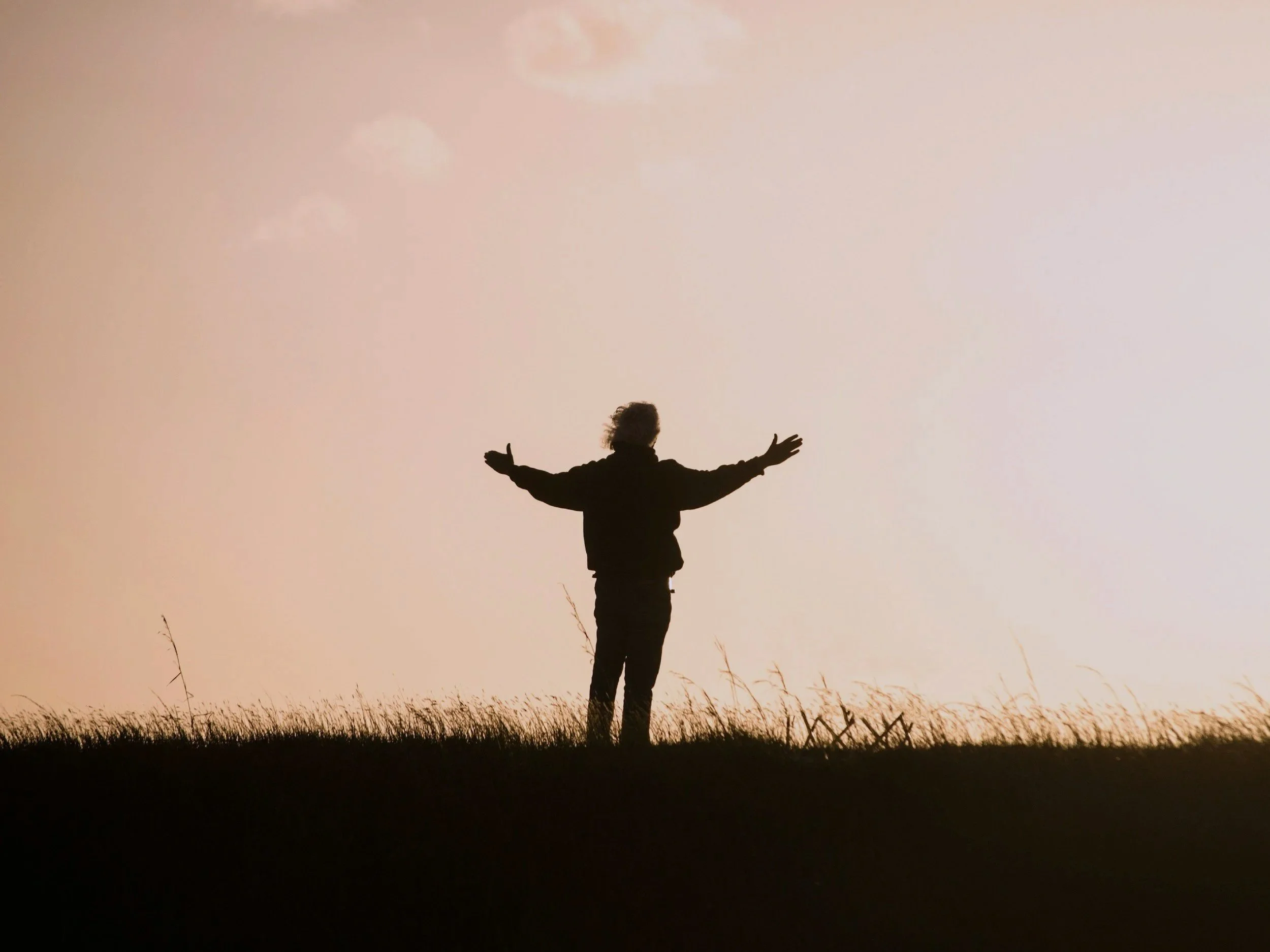 Silhouette of a person standing on grass with arms outstretched, facing the sky during sunset or sunrise.