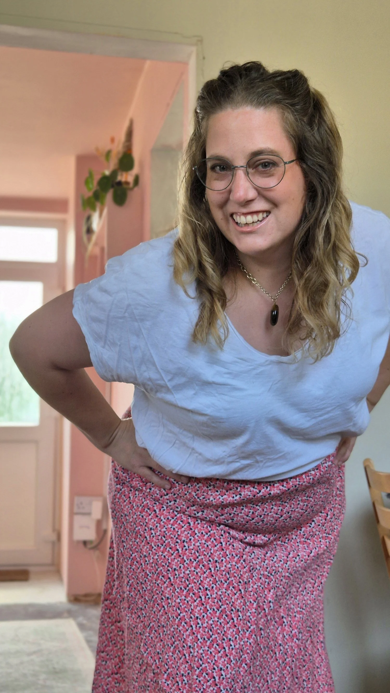 Woman with glasses smiling, wearing a white top and pink patterned skirt, standing in a cozy, well-lit kitchen.
