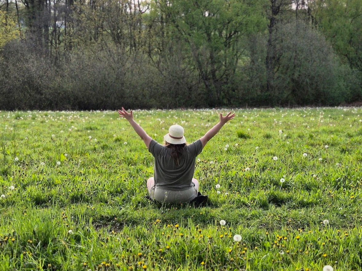 Person sitting on a grassy field with their back to the camera, arms raised, wearing a hat, with trees in the background.