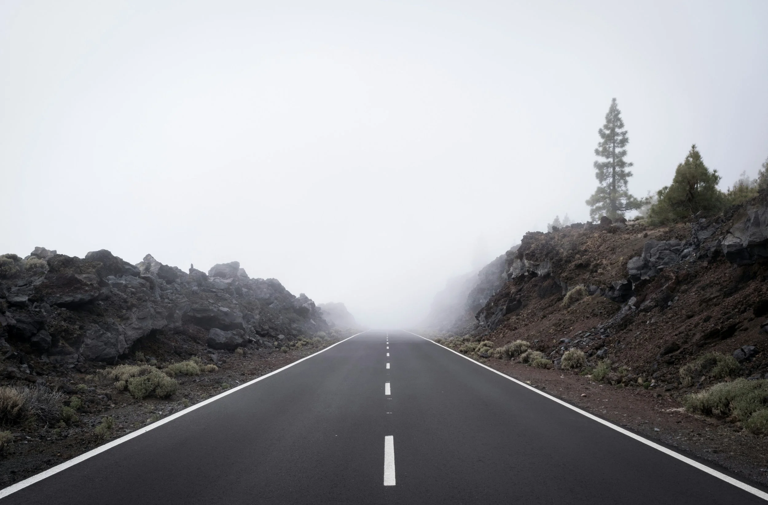 Une route désertique qui traverse une zone volcanique avec des roches noires et des collines, entourée de brouillard ou de nuages, avec quelques sapins en arrière-plan.