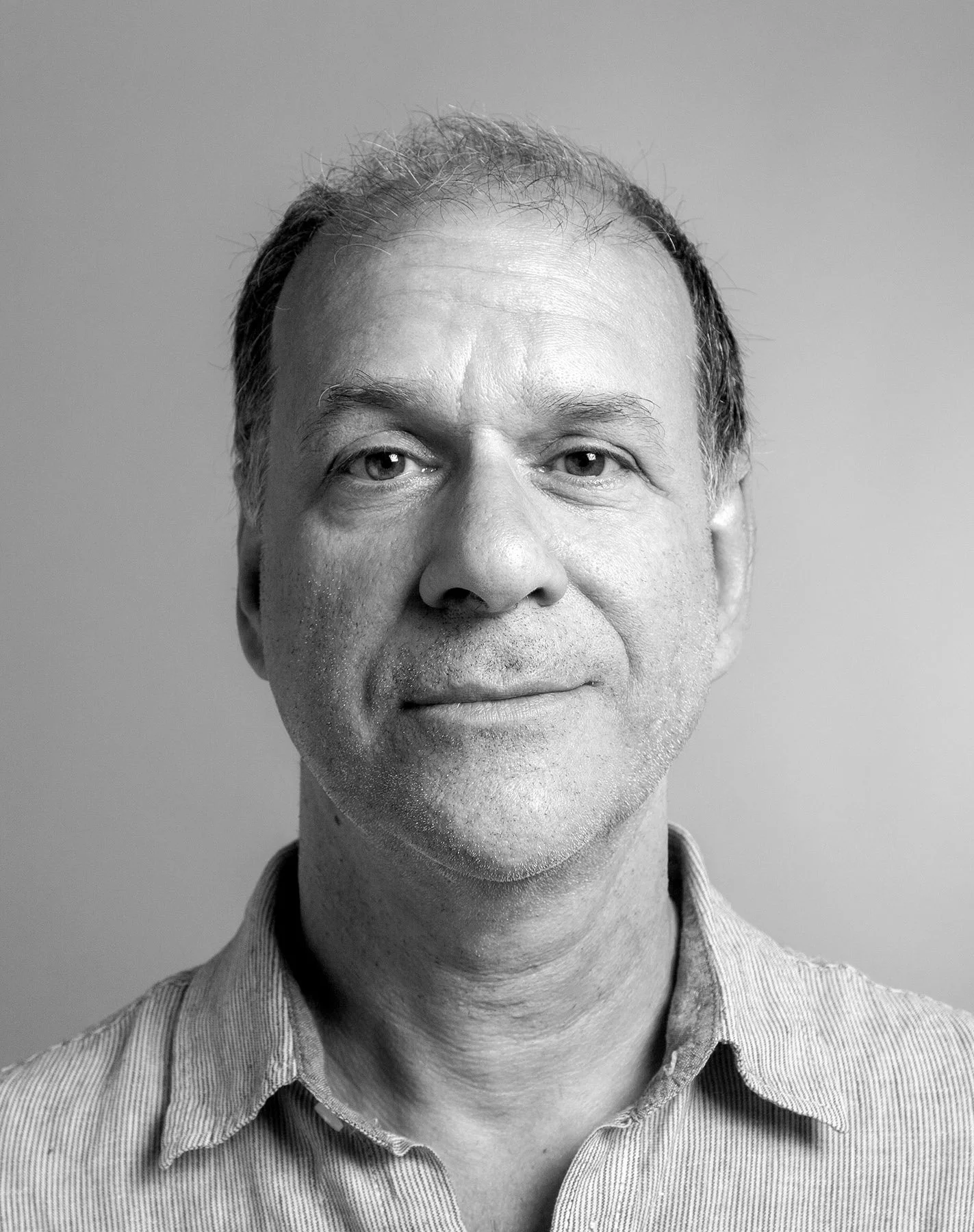 Photo en noir et blanc d'un homme avec des cheveux courts, portant une chemise à rayures, souriant légèrement, face à l'appareil.