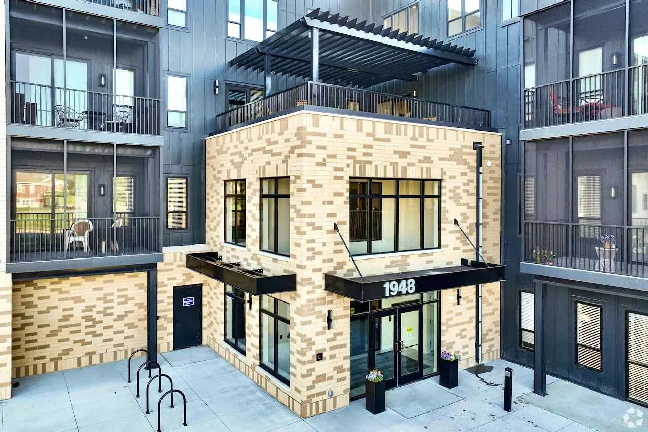 A modern apartment building with a brick facade on the ground floor and black siding on the upper floors. There are balconies with outdoor furniture on each level, and a covered rooftop terrace. The building's address, 1948, is displayed above the entrance.