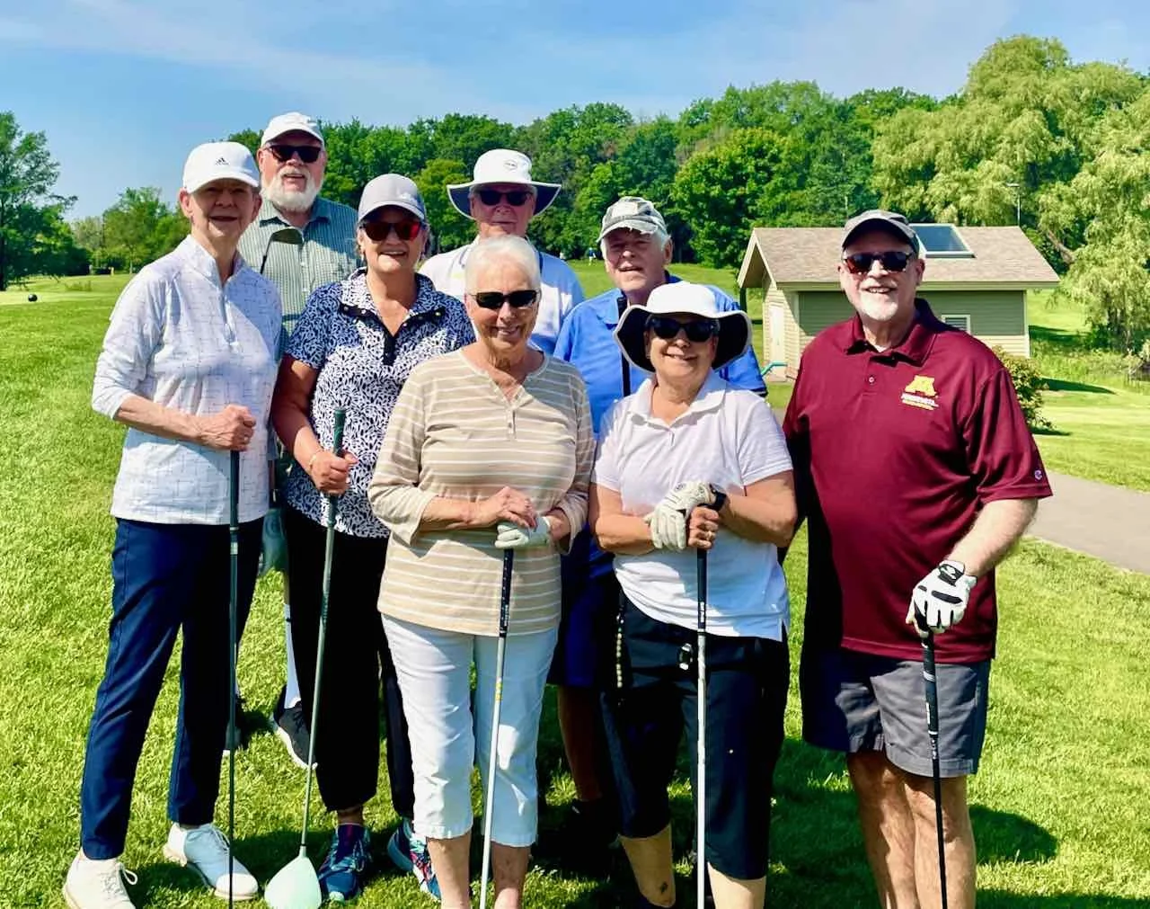 Group of eight older adults standing on a golf course, holding golf clubs, smiling, with green trees, a small building, and a bright blue sky in the background.