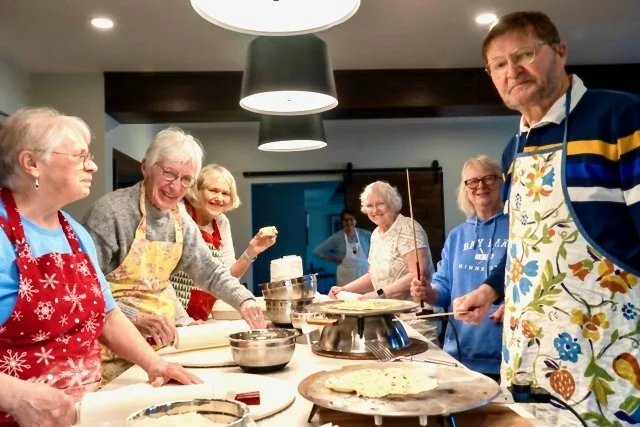 A group of older women and a man in a kitchen making pizza dough, smiling and wearing aprons.