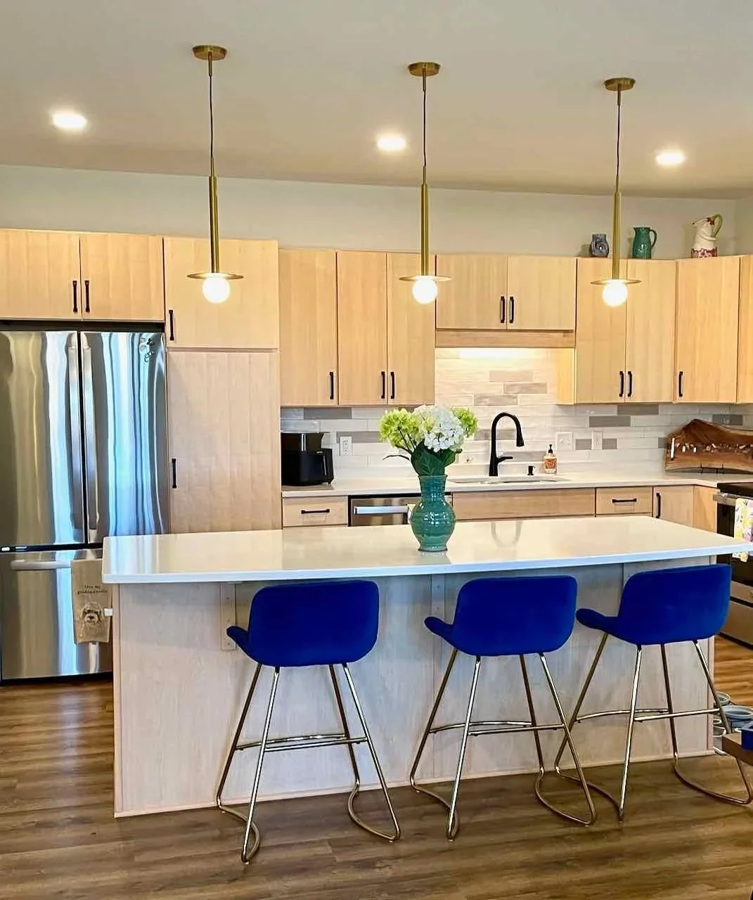 Modern kitchen with wooden cabinets, stainless steel refrigerator, black faucet, white countertop, three blue bar stools, a green vase with white flowers, and pendant lights.