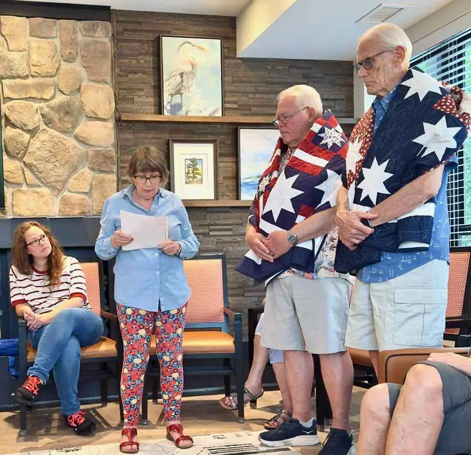 A woman reads from a paper in a room with stone and wood wall decor, while two men stand in front of her, two wrapped in patriotic quilts. A young woman with long hair and glasses sits nearby, listening.