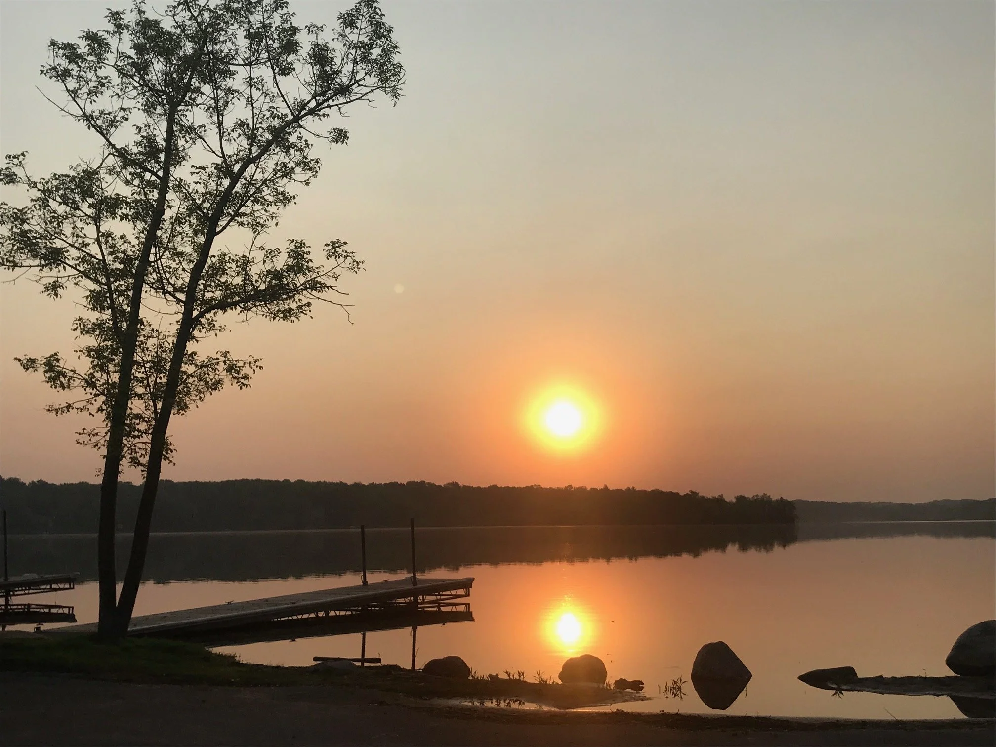 Sunset over a calm lake with a dock, rocks along the shore, and a tree on the left side.