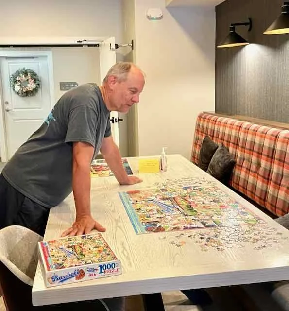 An older man in a gray shirt leans on a large table, working on assembling a 1000-piece puzzle. The table has a partially completed puzzle on it, with a puzzle box nearby. The room has a cozy dining area with a bench seat against the wall, decorated with pillows and a plaid cushion, and a wall with black light fixtures.