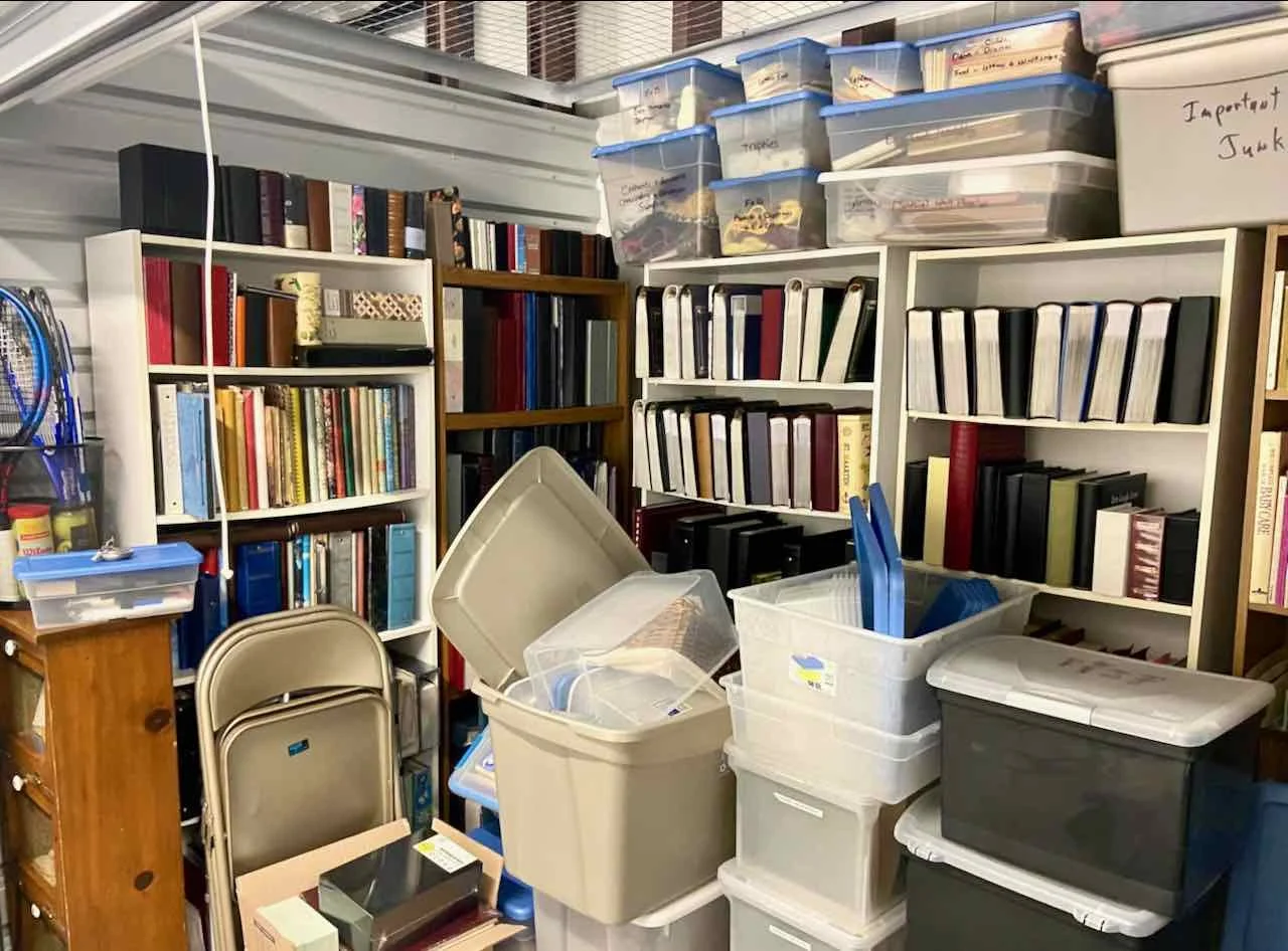 A storage room with white bookshelves filled with binders and books, multiple plastic storage bins on the floor, some open, with folders and papers inside, and a whiteboard with handwritten notes.