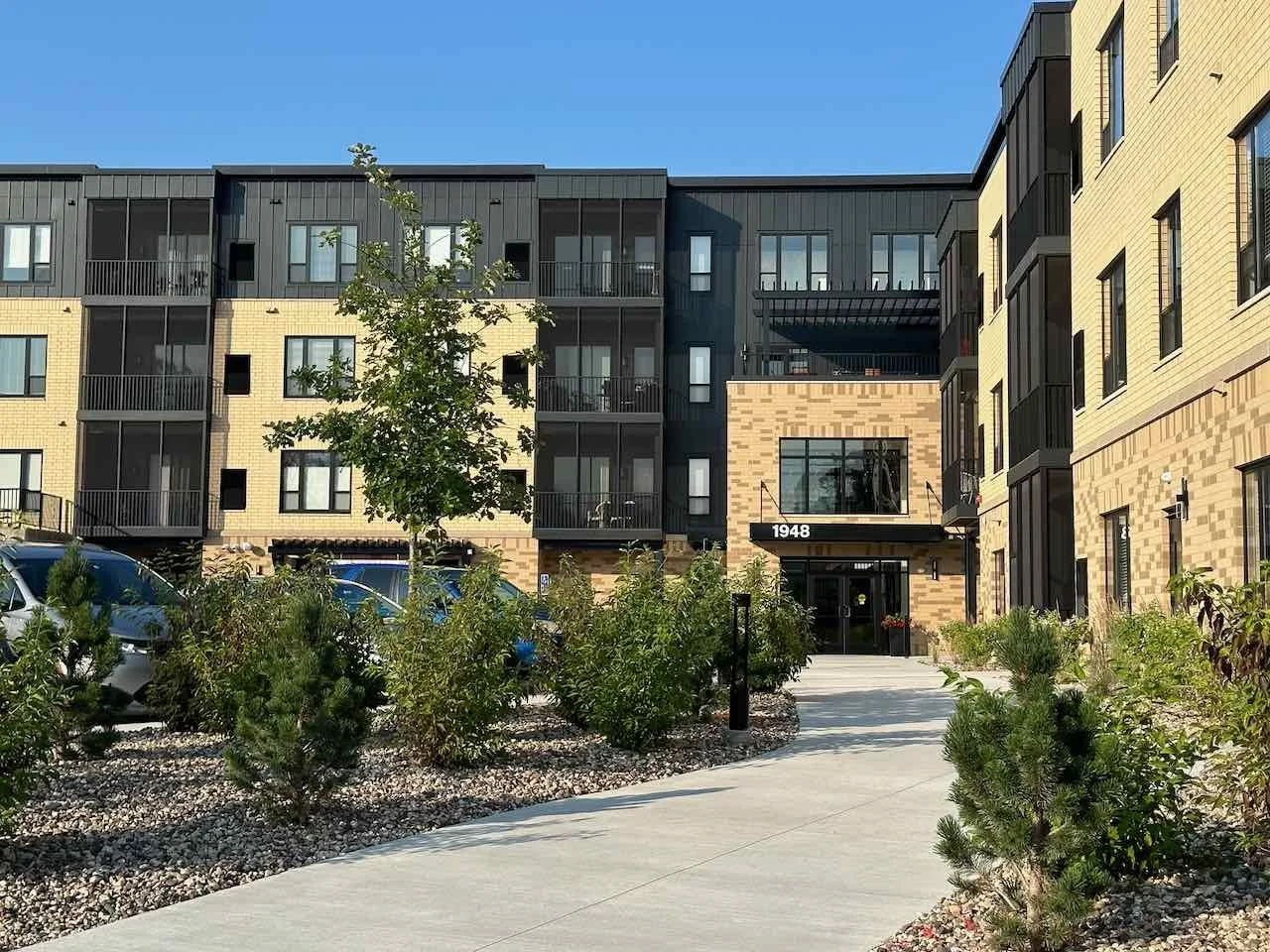 Exterior view of a modern apartment complex with a landscaped walkway, young trees, and parked cars, under a clear blue sky.
