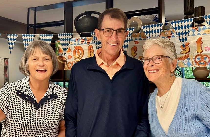 Three smiling adults, two women and one man, standing indoors in front of a decorative backdrop with blue and white patterned bunting, celebrating a birthday.
