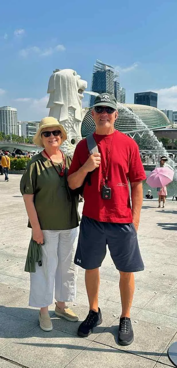 A smiling man and woman posing in front of the Merlion statue in Singapore, with modern city buildings in the background. The woman wears a hat and sunglasses, while the man wears a cap and sunglasses. They're dressed casually, suitable for sightseeing.