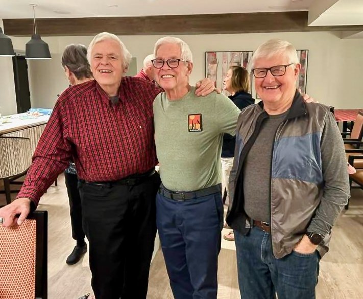 Three older men smiling and standing together at an indoor gathering, with other people in the background.