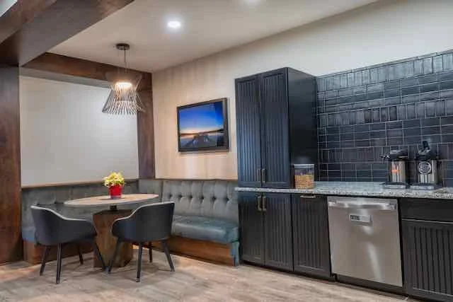 A kitchen and dining area with a table, four chairs, a banquette, a black cabinet, and kitchen appliances. There is a television on the wall and a decorative light fixture hanging from the ceiling.