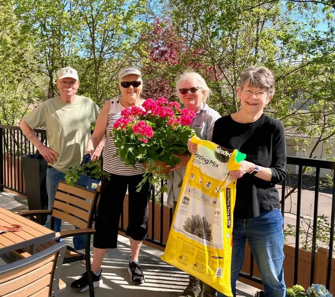 Four smiling women and one man standing on a balcony with trees behind them, holding a potted pink flowering plant and a bag of soil or gardening mix.