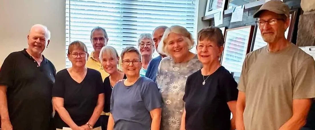 Group of ten smiling older adults standing indoors near a window with blinds and framed pictures on the wall.