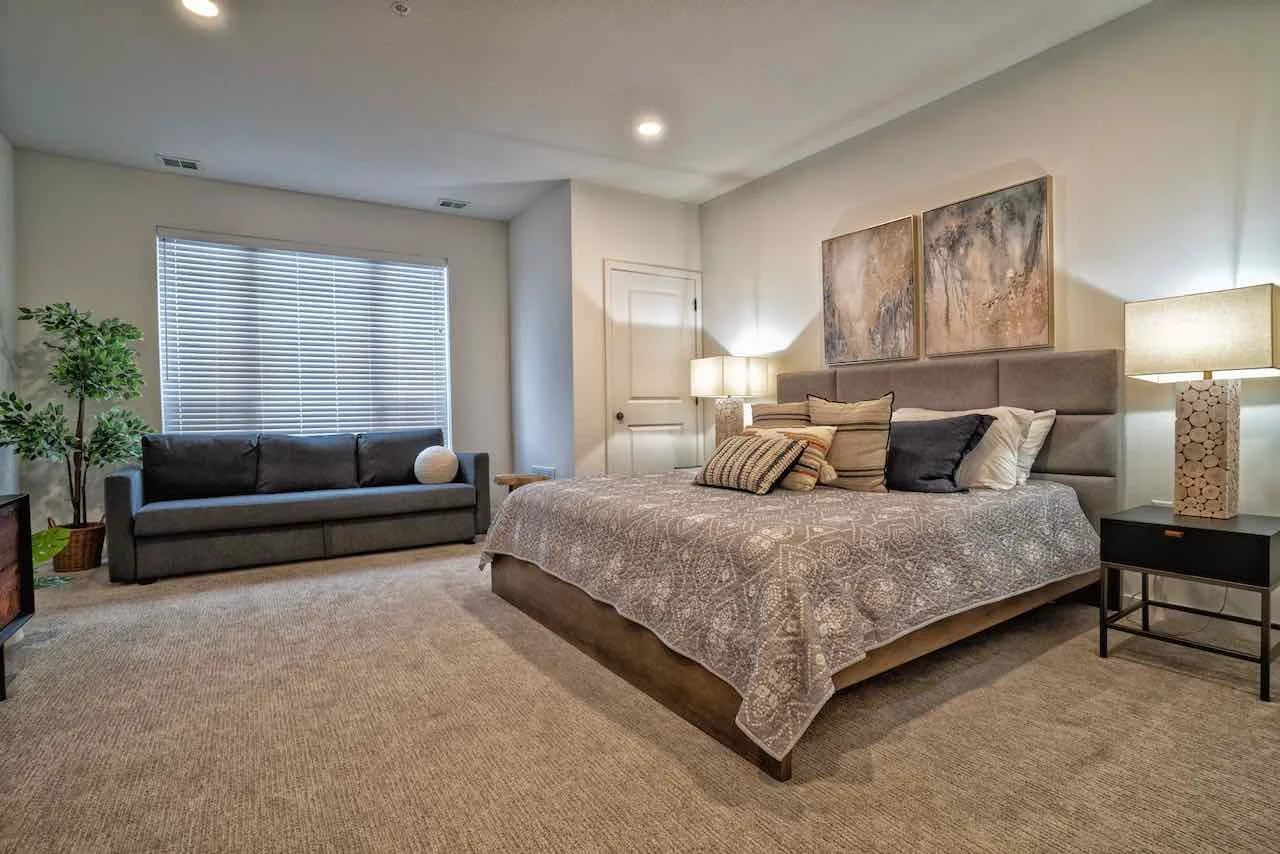 Bedroom with king-sized bed, gray headboard, patterned bedding, and multiple pillows, two bedside lamps, window with blinds, gray sofa with a round pillow, indoor plant, and artwork on the wall.