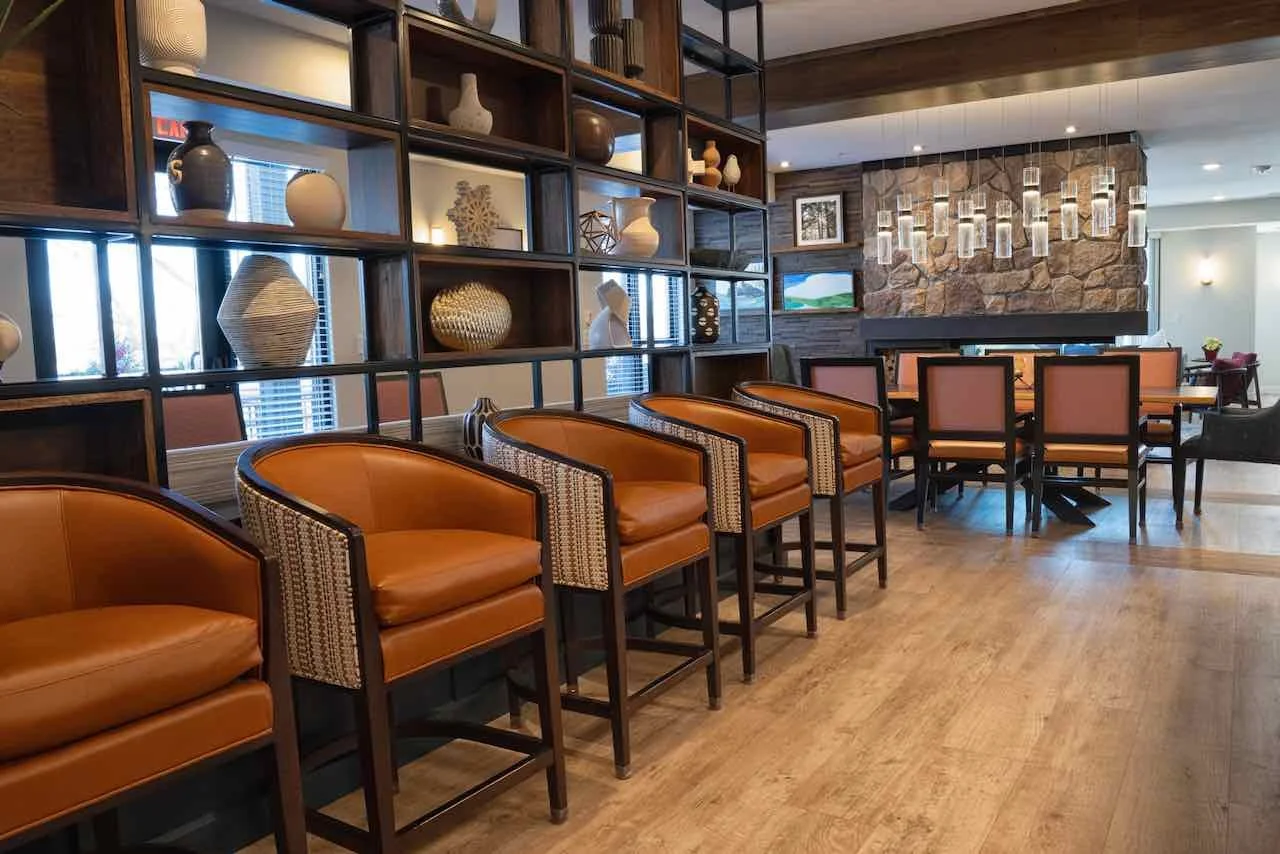 Interior of a lounge with a wooden floor, a dividing shelf with decorative vases and sculptures, a stone fireplace with hanging lights, and seating including leather and upholstered chairs around tables.