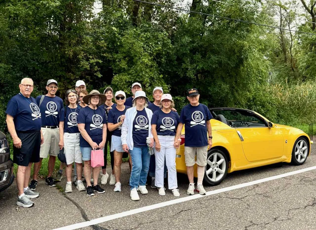 Group of older adults in matching navy blue shirts standing in front of a yellow convertible car on a wooded roadside.