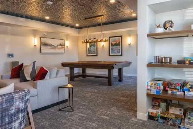 A living room with a pool table, framed landscape art on the wall, and a built-in bookshelf filled with books and decorative items.