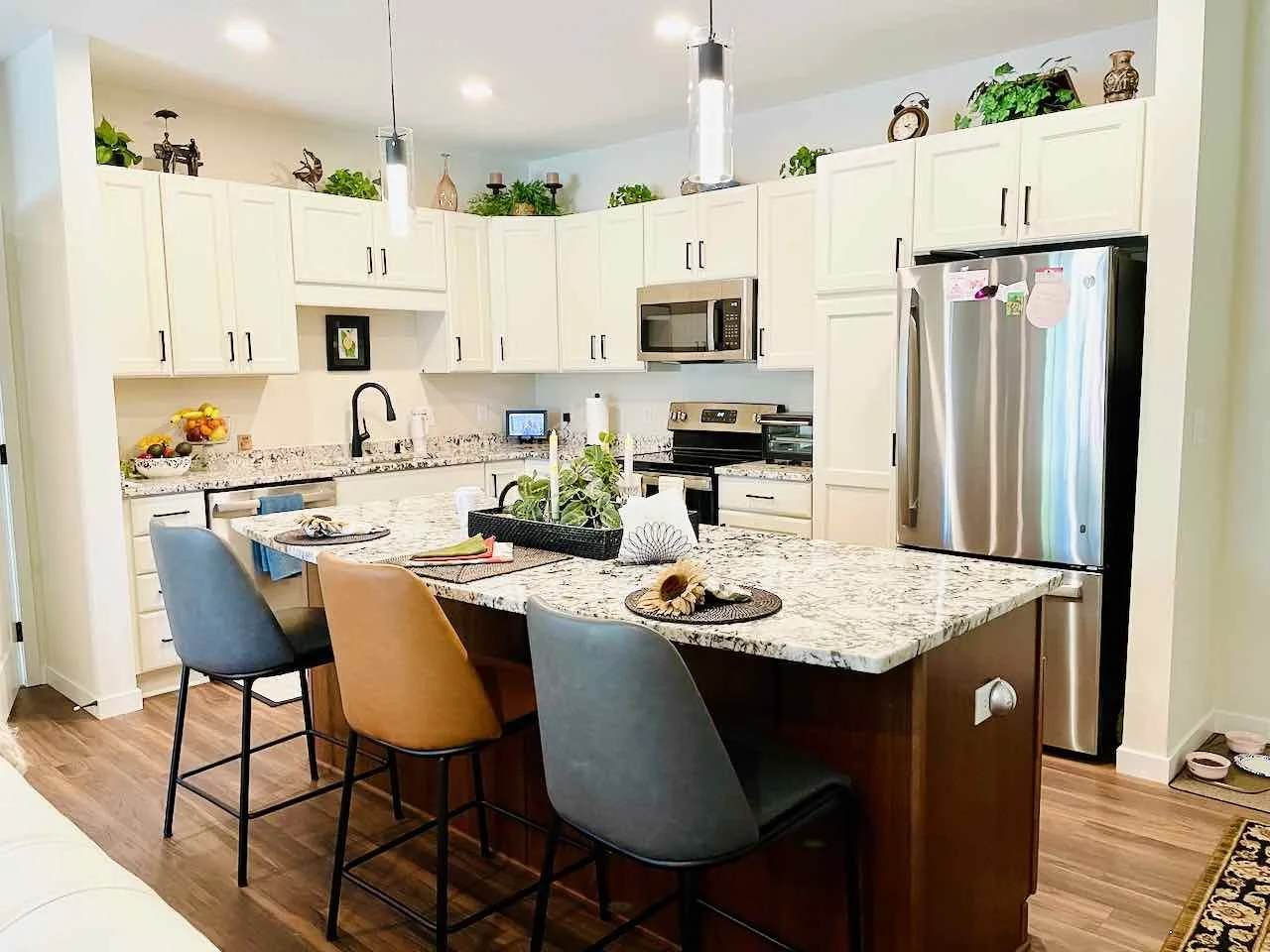 Modern kitchen with white cabinets, stainless steel refrigerator, microwave, and granite countertops. There are colorful chairs around a kitchen island with decorative items and plants on top.