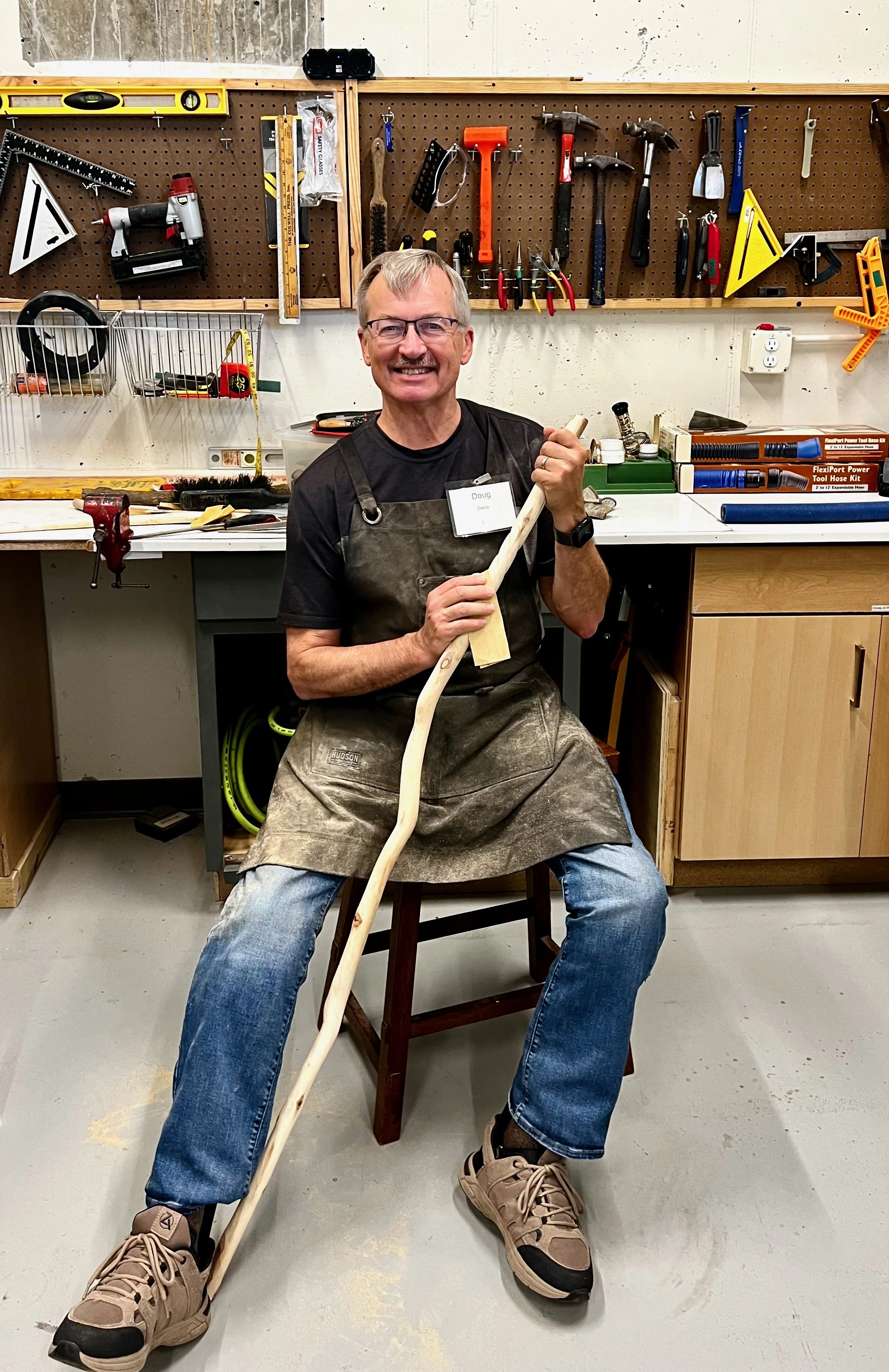 A man sitting on a stool in a woodworking workshop, smiling, holding a wooden cane or staff, wearing a black T-shirt, glasses, apron, and sneakers. The workshop has various tools hanging on a pegboard behind him and a workbench with supplies and tools.