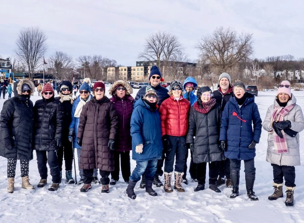 A group of about fifteen people dressed in winter clothing standing on a frozen lake with during winter. They are smiling and posing for the photo with trees, buildings, and cars in the background.