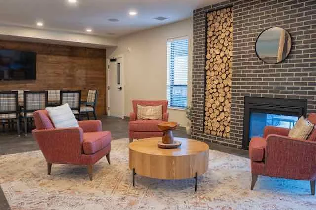 Living room with red armchairs around a round wooden coffee table, a fireplace, a window, and a wall of firewood logs.