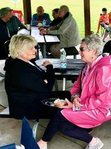 Two older women sitting and talking at a picnic table outdoors. One woman is in a black jacket, and the other is in a pink jacket holding a plate with food. Other people are sitting at tables in the background under a pavilion.