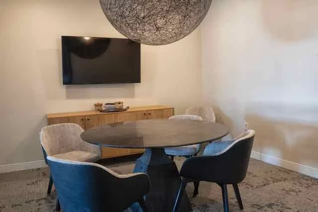 A modern dining room with a round table surrounded by four upholstered chairs, a large spherical pendant light overhead, a flat-screen TV on the wall, and a wooden sideboard beneath the TV.