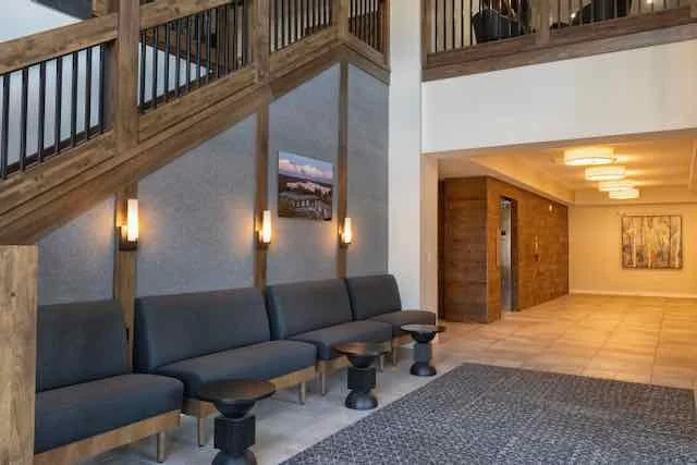 Interior of a building's lobby or waiting area with a row of five dark gray chairs, small black tables, wooden accents, a staircase with wooden railing, and warm lighting.