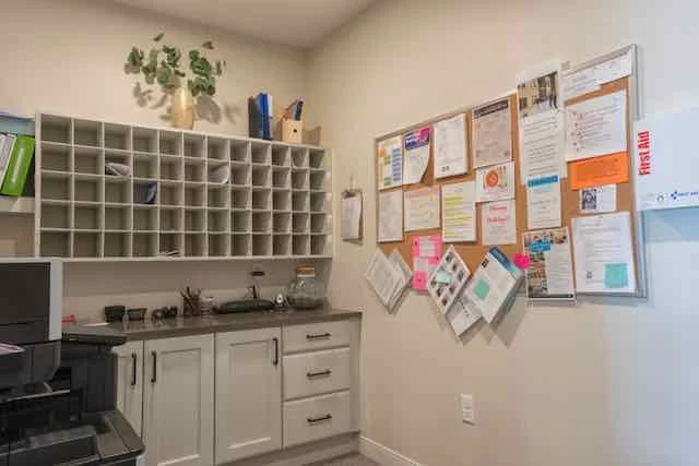 Office kitchenette with a corkboard on the wall filled with notices, a small sink, shelves with mailboxes, and various items on the countertop.