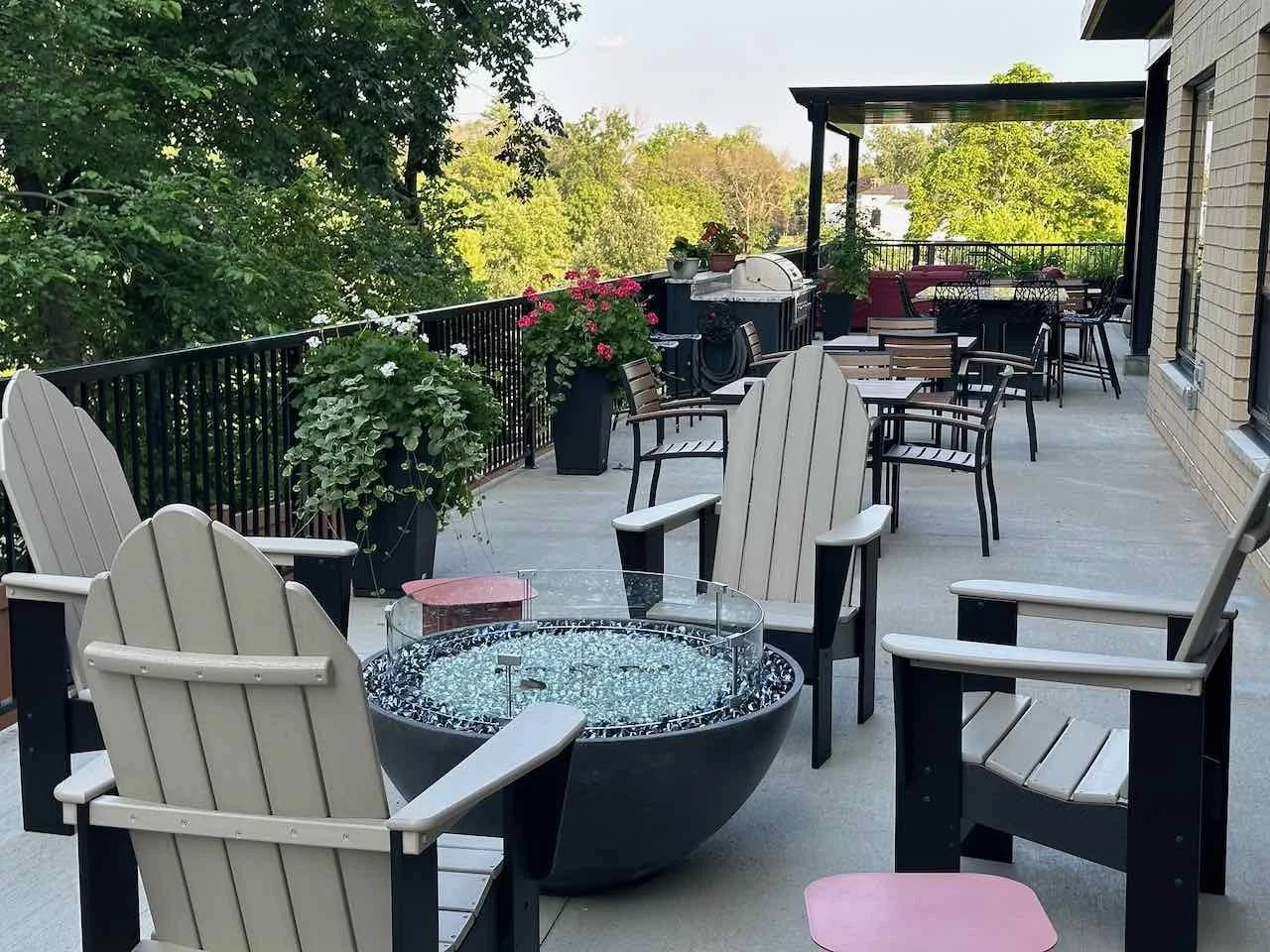 An outdoor patio on a balcony with various seating options, potted flowers, a fire bowl, a barbecue grill, and a small bar area, surrounded by green trees.