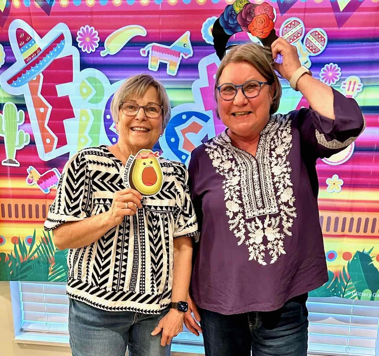 Two women standing in front of a colorful, festive backdrop with Mexican-themed decorations including sombreros, cacti, and maracas. One woman is holding a sign with a surprised avocado character, and both women are smiling, wearing glasses, and casual clothing.