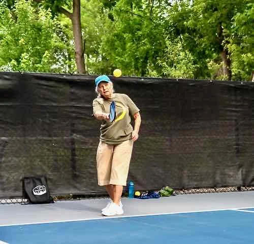 A woman on a pickleball court hitting a pickleball with a racket. She is wearing a blue cap, beige shorts, a beige shirt, and white shoes. There are trees in the background and a black privacy fence behind her.