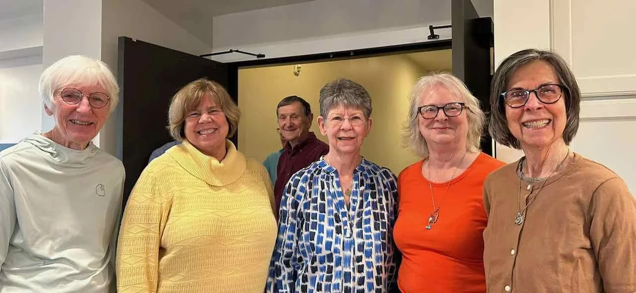 Six smiling older adults, five women and one man, standing together in front of a yellow screen, indoors.