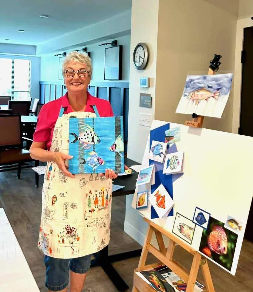 A woman with glasses, short curly gray hair, wearing a pink shirt and a light-colored apron, standing in a room with artwork, including paintings of fish, holding a painting of a fish in front of her.