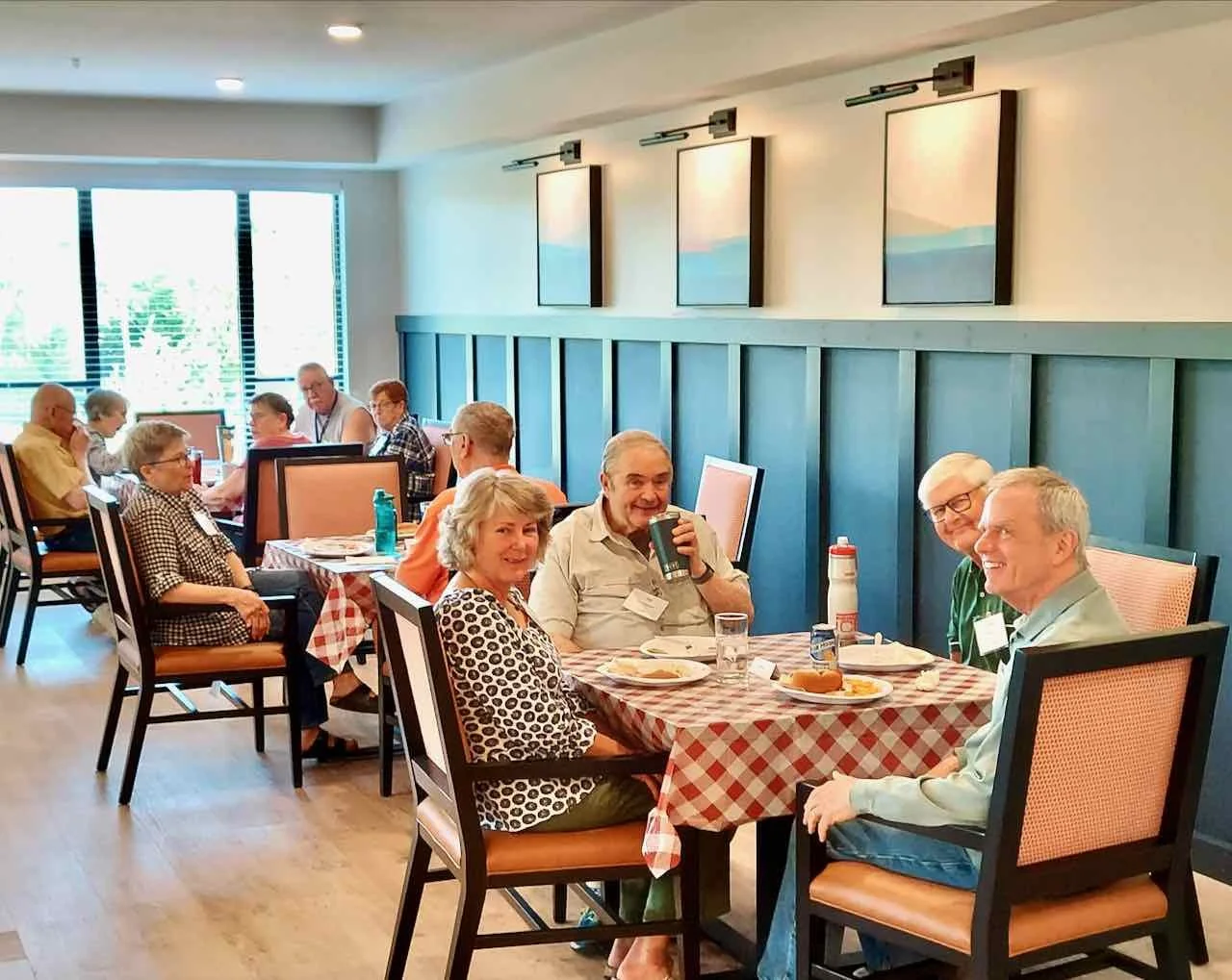 A group of older adults sitting around two tables in a bright restaurant or banquet room. They are smiling and enjoying a meal together, with some looking towards the camera. The room features large windows, blue wall paneling, and artwork on the walls.