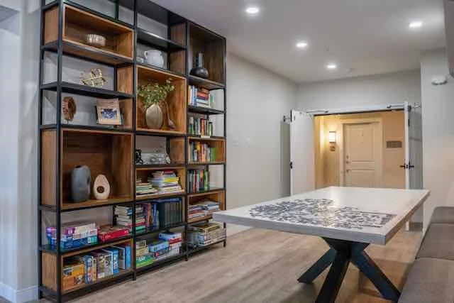 A room with a large wooden bookshelf, a large white table with a black base, a beige sectional sofa, and a door leading to another room. The room has hardwood floors and recessed ceiling lights.