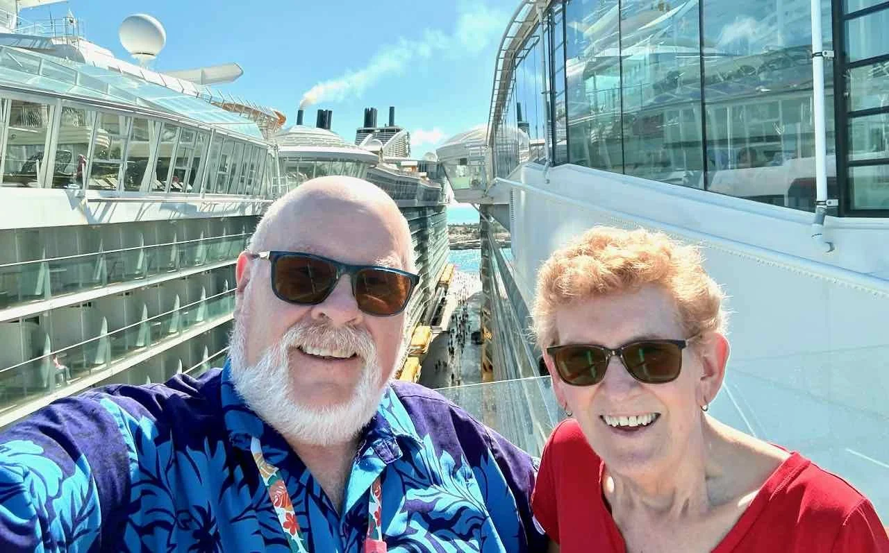 Smiling man with white beard and woman wearing sunglasses taking a selfie on a cruise ship balcony with other cruise ships docked nearby under a bright blue sky.