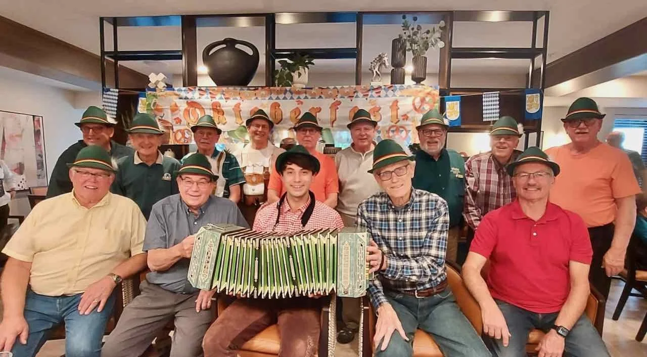 A group of  men and women dressed in traditional Bavarian attire, wearing green hats with feathers, are gathered around in a restaurant or pub, posing for a photo with a young man holding an accordion. The background has a decorative banner and shelves with vases and plants