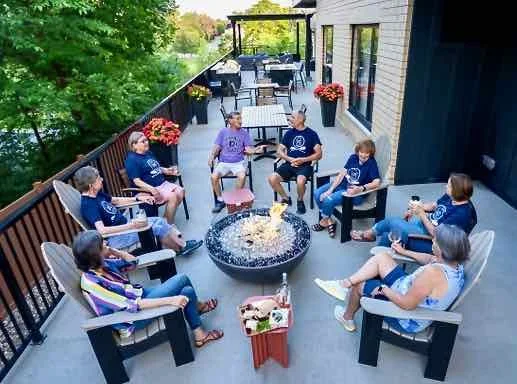 Group of people sitting around a fire pit on a balcony, enjoying conversation and beverages, with tables, chairs, and potted plants in the background.