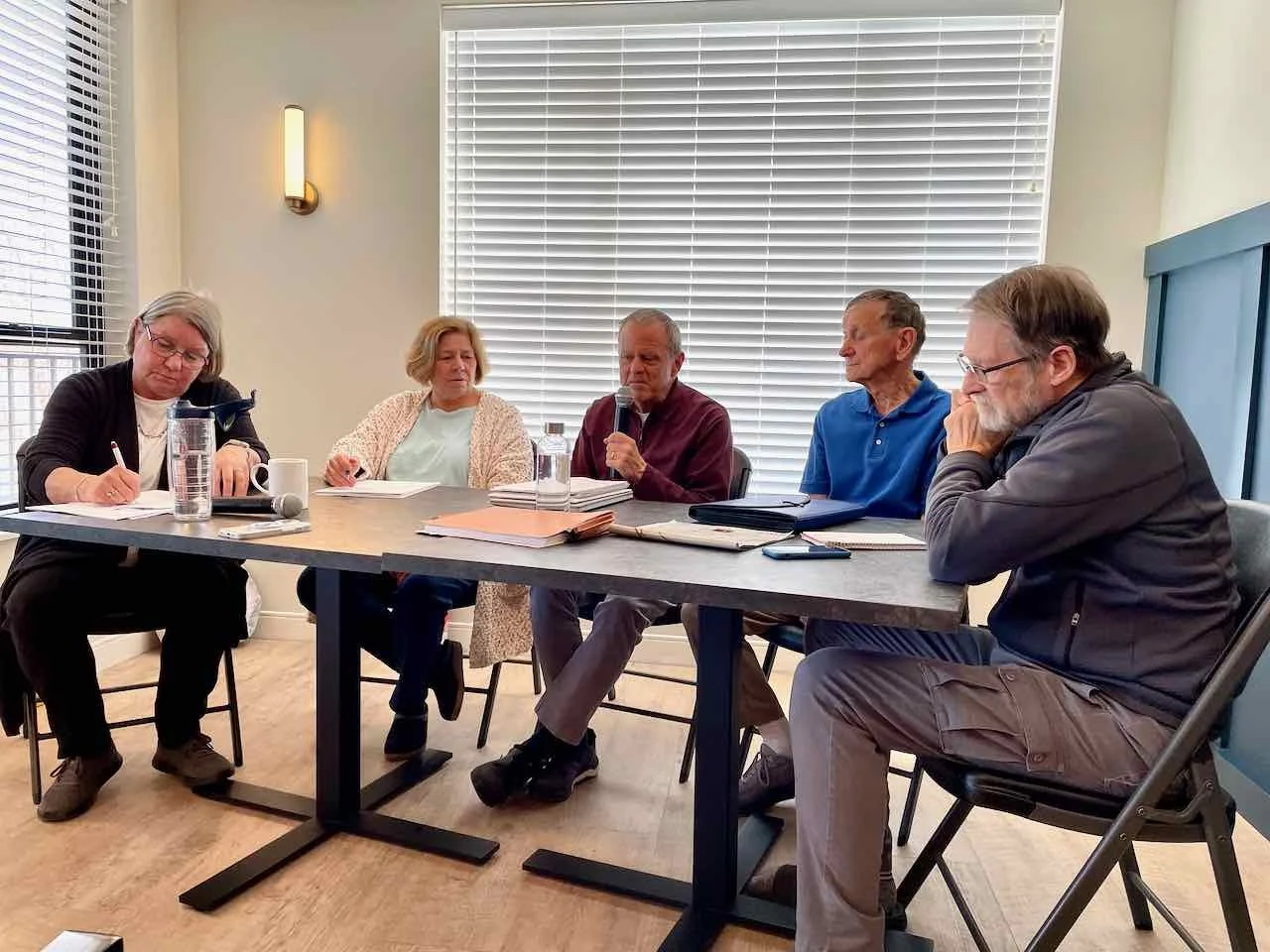 Five adults seated at a conference table engaged in a meeting, with papers, notebooks, a water bottle, a mug, and a microphone on the table, in a room with large window blinds and a wall light.