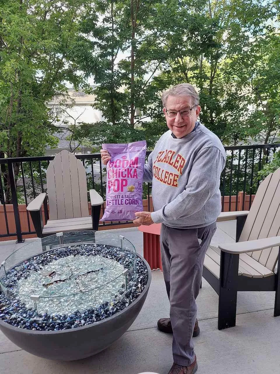 A man is standing outdoors on a deck, holding a bag of Wounded Chicka Pop kettle corn. He is smiling and wearing glasses, a gray sweatshirt, and gray pants. There are chairs and trees in the background, and a fire pit with a glass cover in front of him.