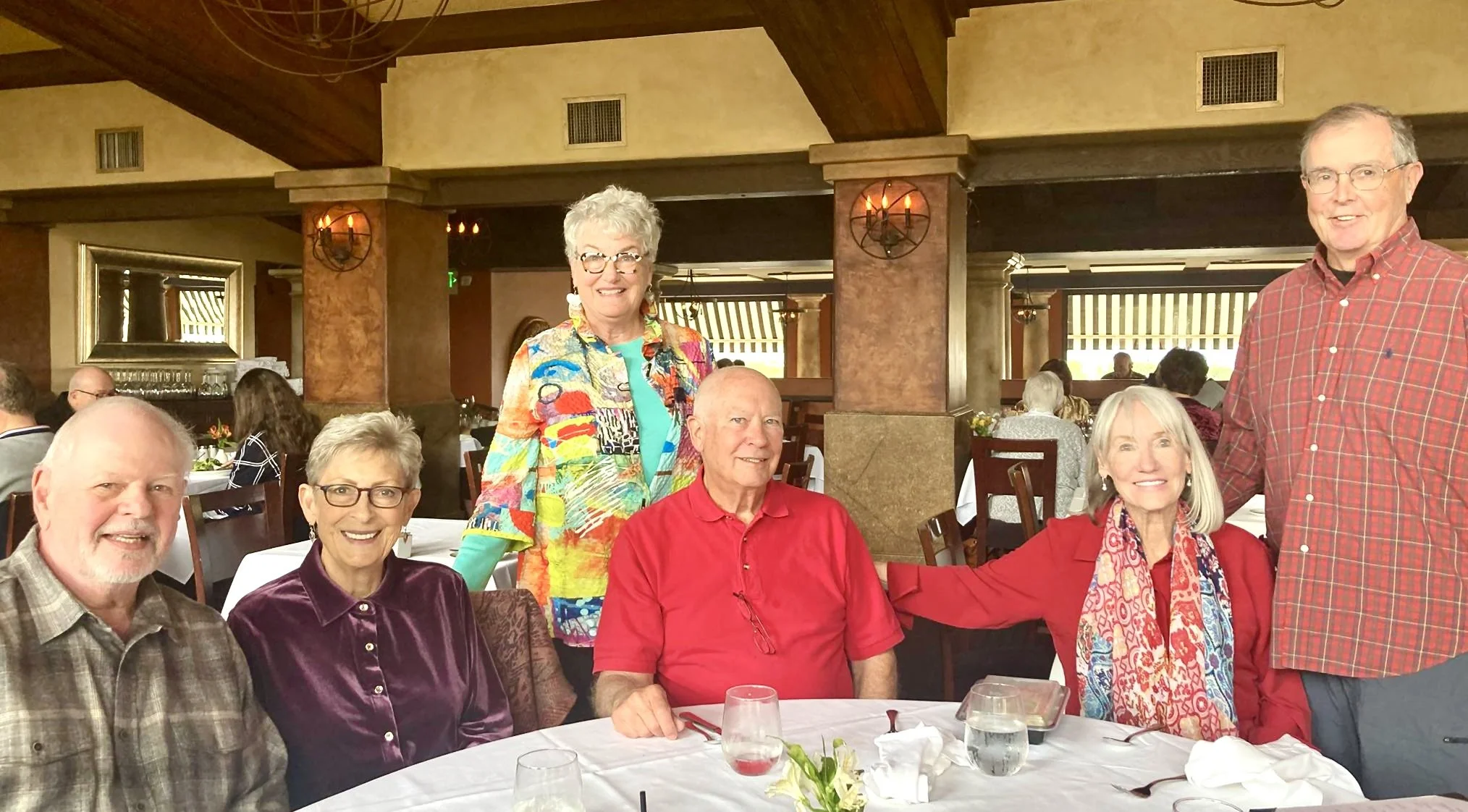 Group of six elderly people, four women and two men, gathered around a table in a restaurant, smiling for the camera. One woman stands behind the seated group, wearing a colorful jacket, while the others are seated in front of her, dressed casually. The table has glasses, napkins, and a small flower arrangement. The restaurant has a warm, inviting ambiance with wood accents and other patrons in the background.