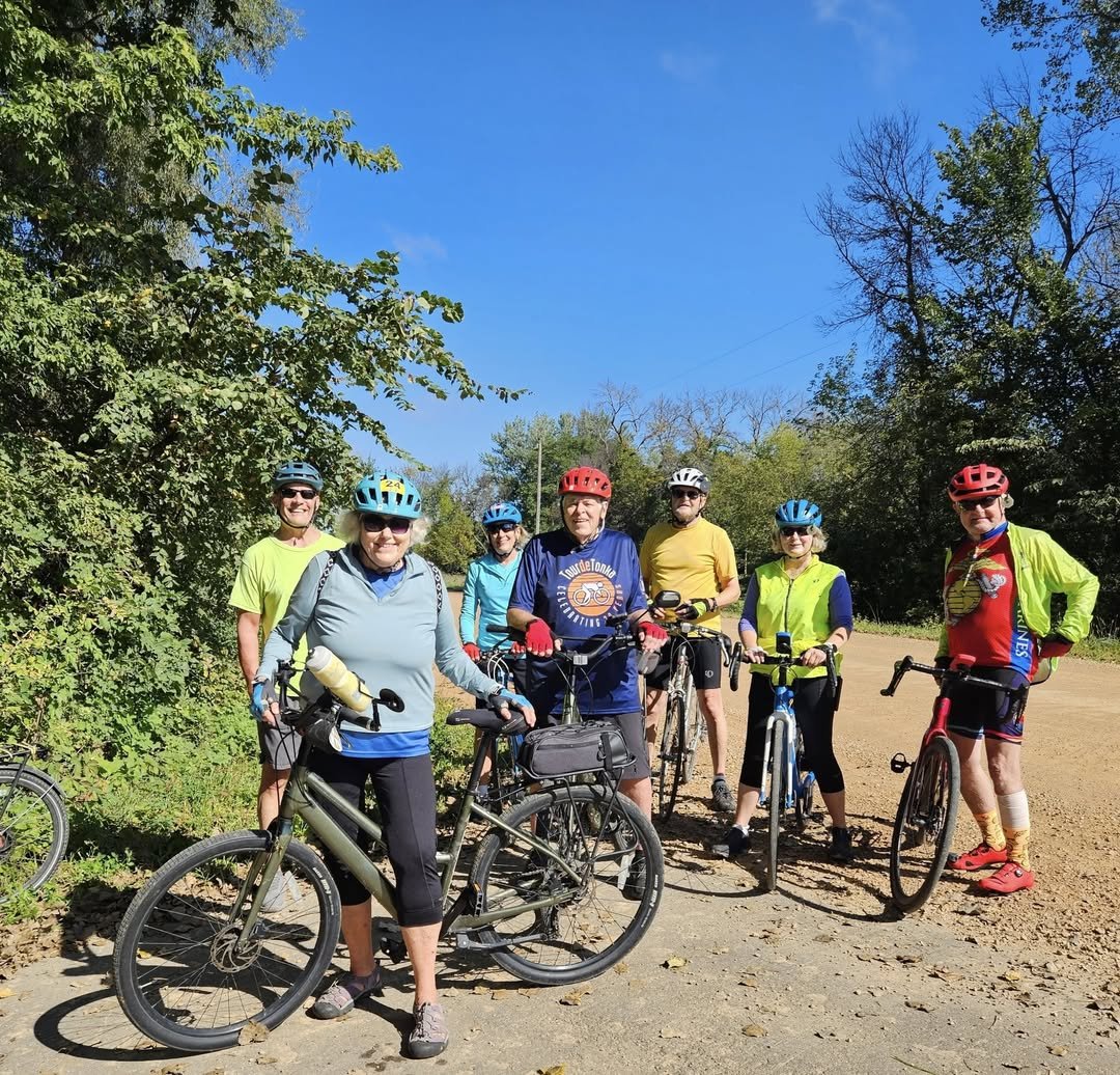 A group of seven bikers wearing bike helmets are standing with their bikes along a bike trail with green trees.