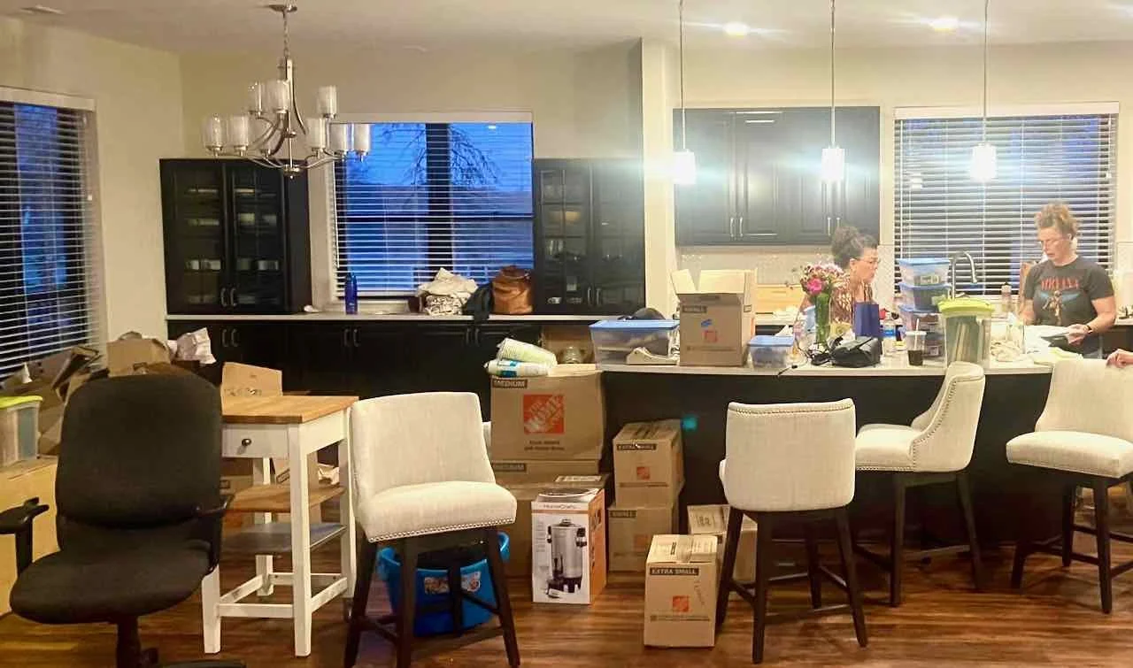 A cluttered kitchen and dining area with boxes and items on the counter and floor, two women standing at the kitchen island, and various chairs around.