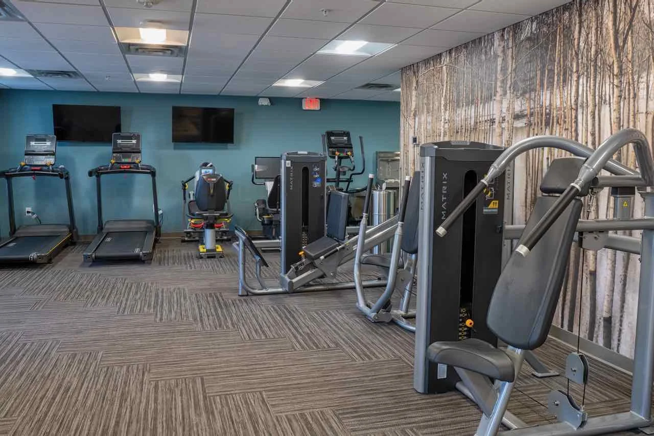 A large, empty gym room with treadmills, stationary bikes, and weight machines, wood paneling on one wall, TV screens, ceiling tiles, and carpeted floor.