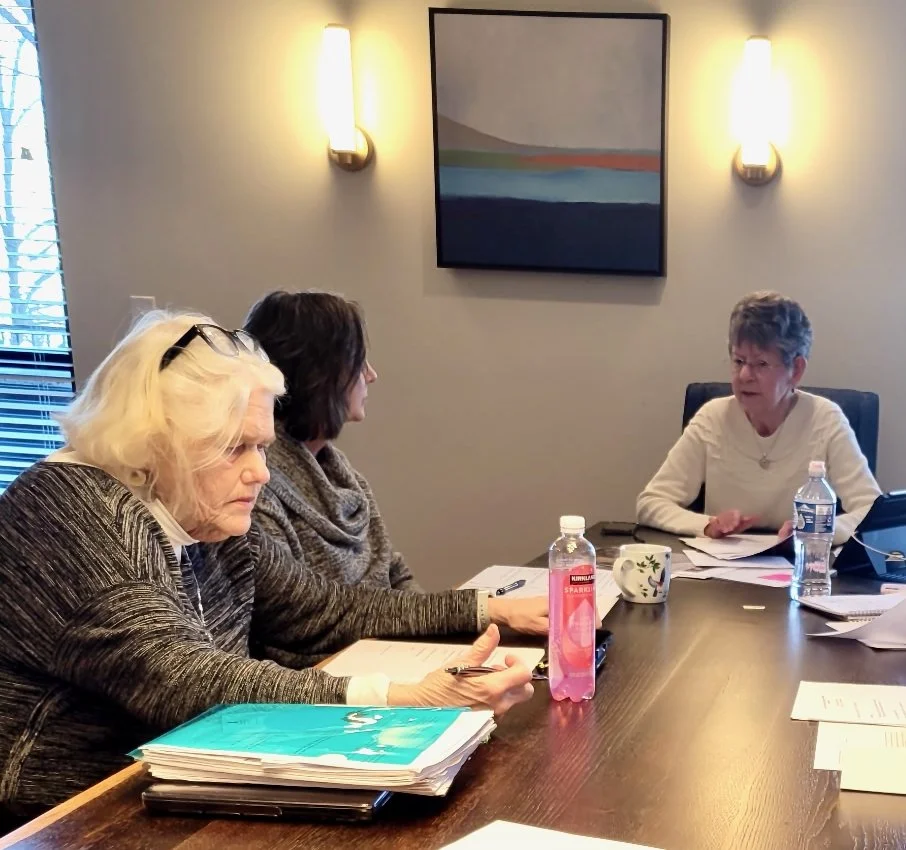 Three women sitting at a conference table in a meeting room, with documents, water bottles, and a mug on the table, engaged in conversation.
